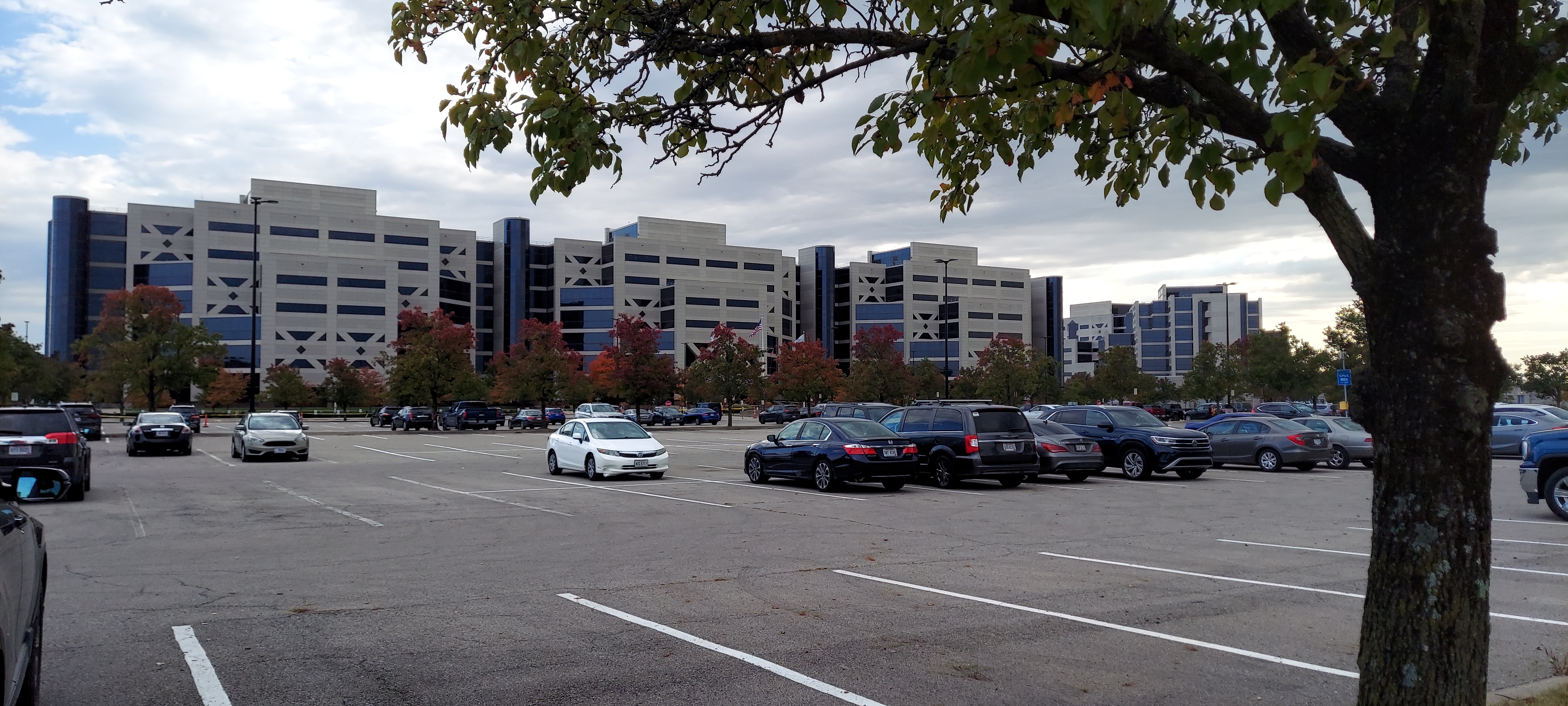 Large building complex which has three sections and is tan with dark windows and rises seven stories. Photo taken in Fall of 2022 which shows the view from the front of the building from the large parking lot. Trees are starting to change colors amidst the cars parked in the lot. 